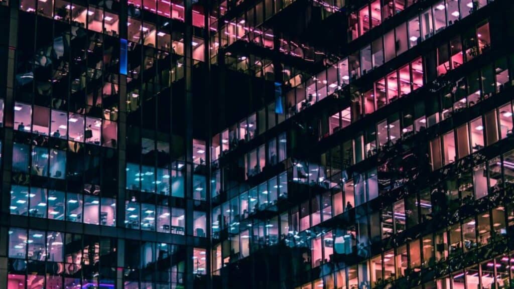 Modern office building at night with many windows lit in contrasting pink and teal colors.