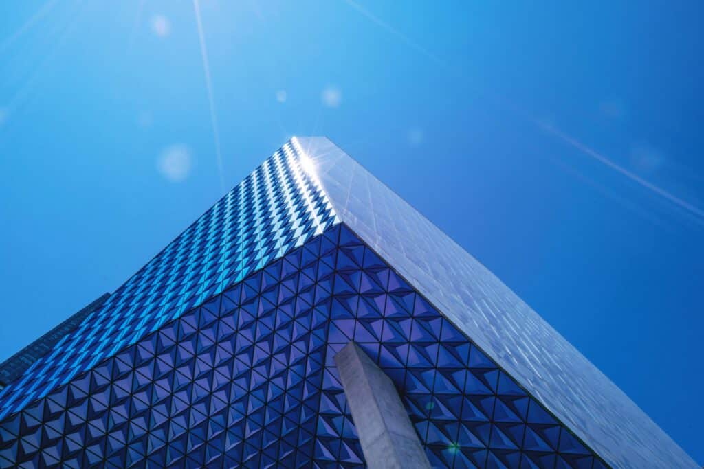 Low-angle view of a modern building with blue and purple geometric panels and reflective glass, under a bright blue sky with sun flares.