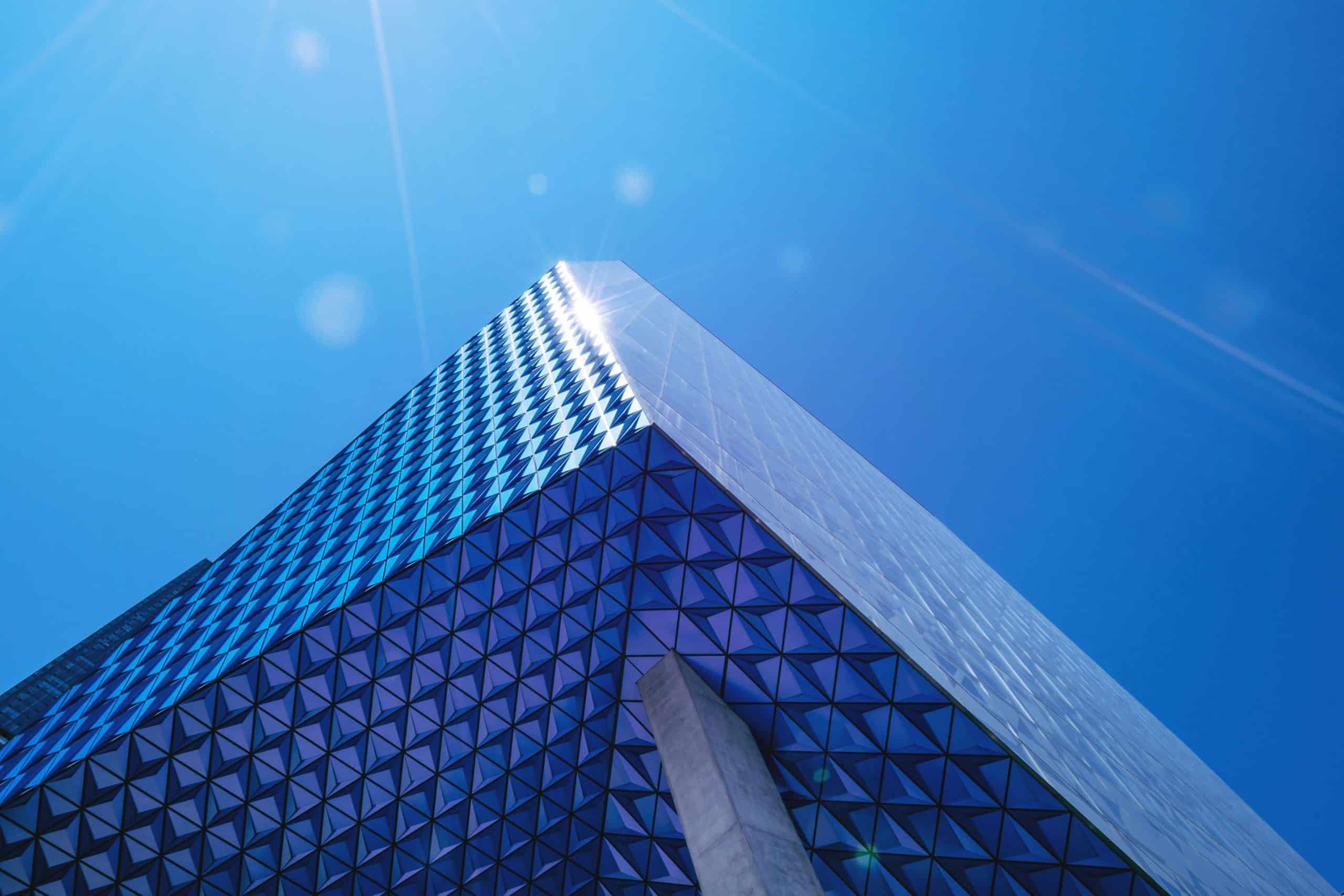 Low-angle view of a modern building with blue and purple geometric panels and reflective glass, under a bright blue sky with sun flares.