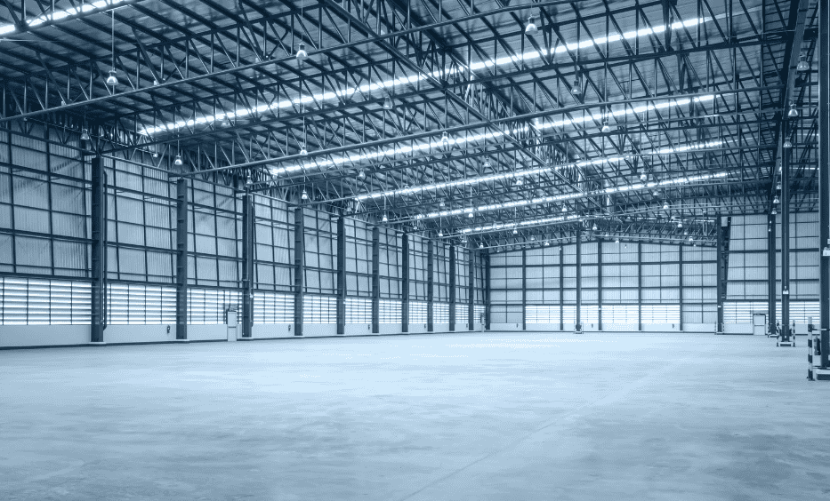 Interior of a large, empty industrial warehouse with a concrete floor, corrugated metal walls, and an exposed metal truss ceiling with bright lighting.