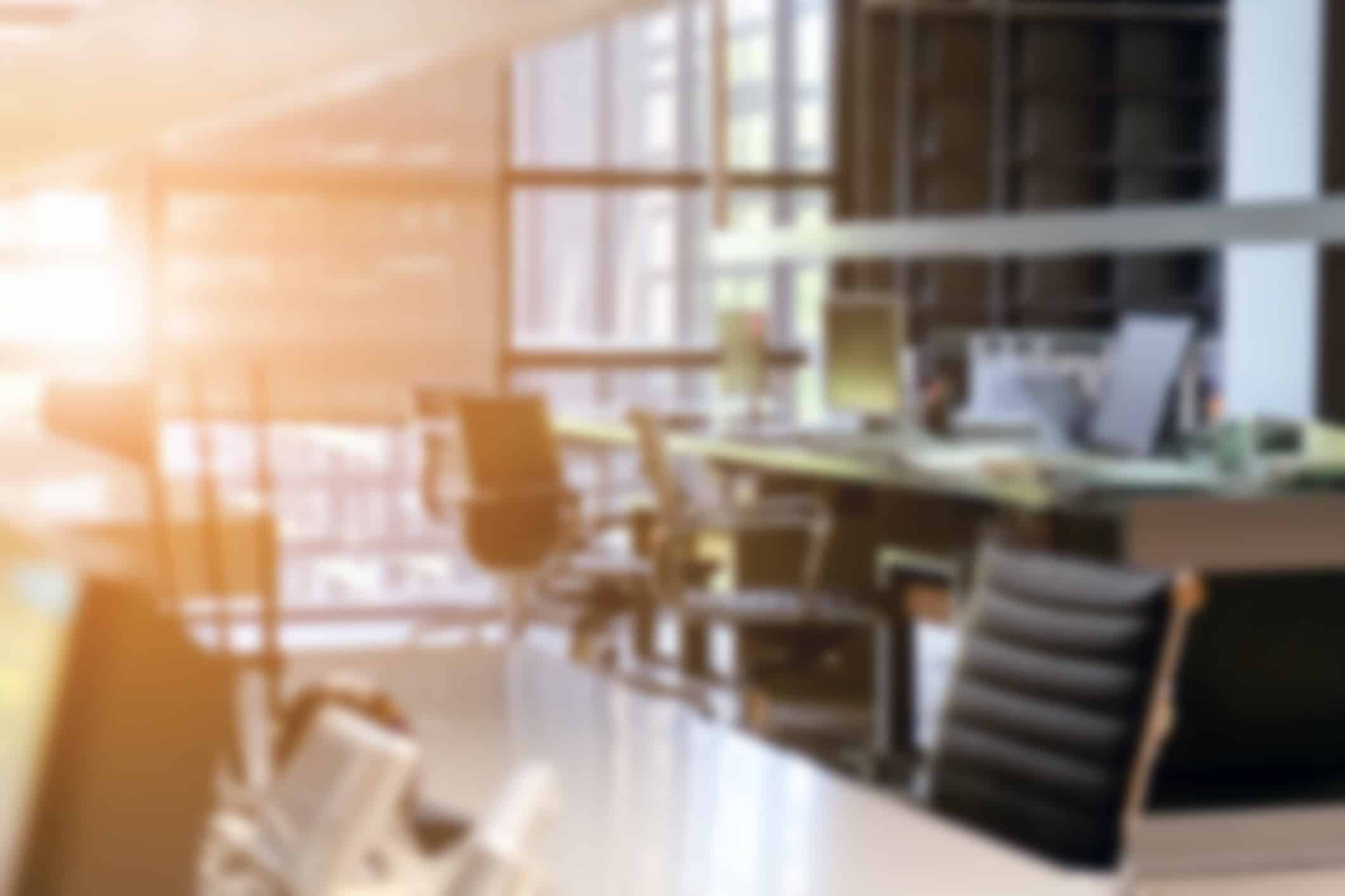 Blurred image of an office interior with desks, chairs, and bright windows with a sun flare.