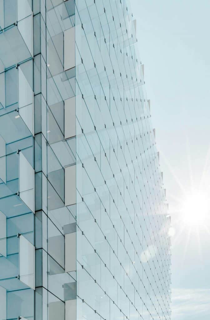 Close-up of a modern building's glass facade with a textured, grid-like pattern of clear and opaque panels, reflecting a bright, sunny sky.