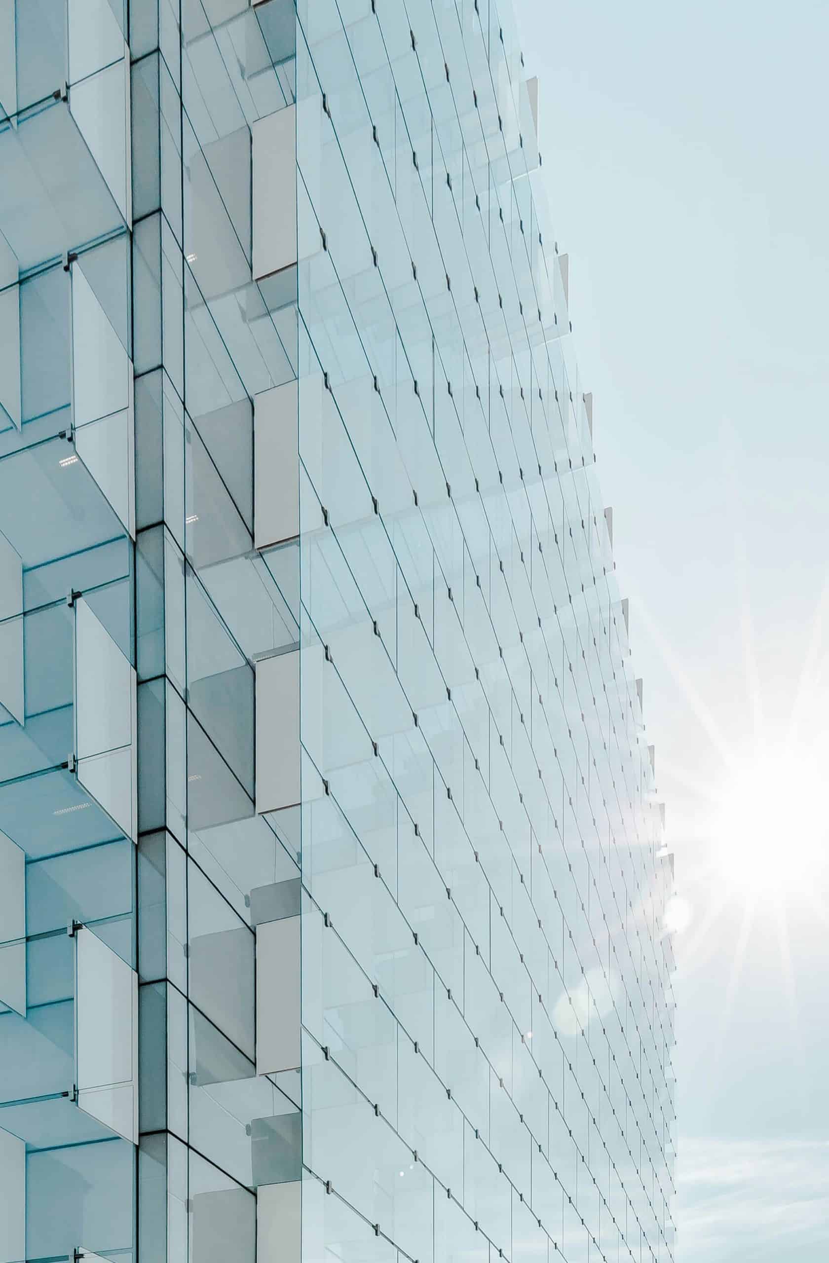 Close-up of a modern building's glass facade with a textured, grid-like pattern of clear and opaque panels, reflecting a bright, sunny sky.
