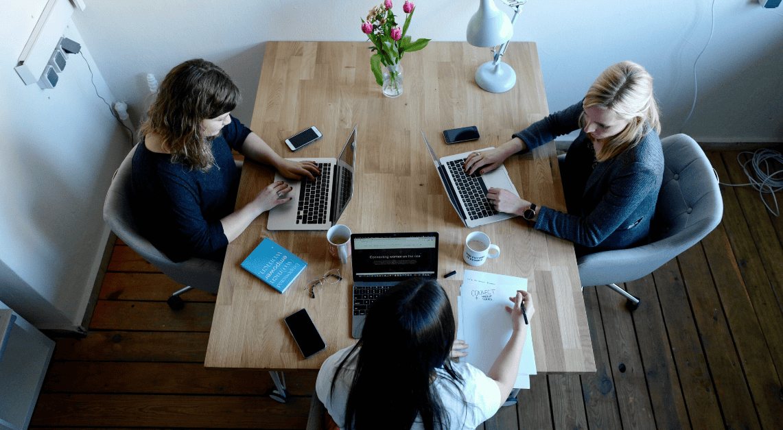 Overhead view of three women working at a wooden table with laptops, notebooks, and coffee mugs.