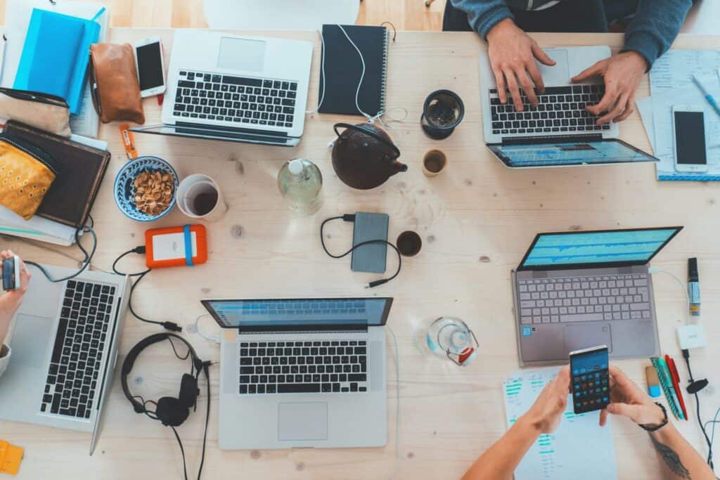 Overhead view of a wooden table with four laptops, multiple smartphones, and hands working, surrounded by notebooks, snacks, and electronic accessories.