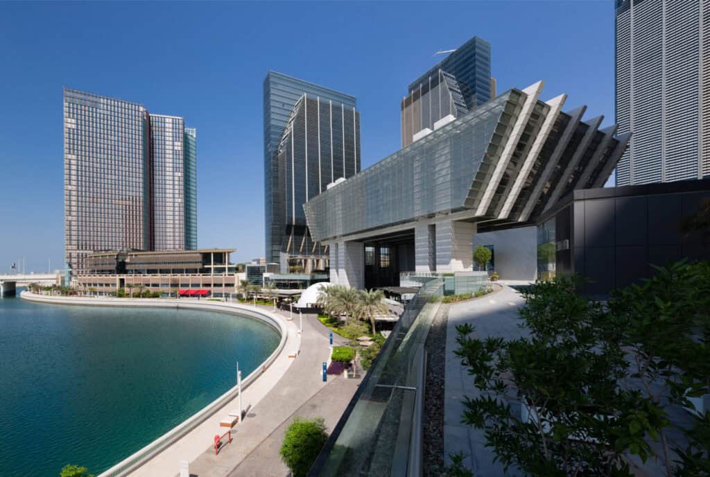 An elevated view of a modern city waterfront with tall glass skyscrapers, a unique cantilevered building, a curved promenade, and a teal body of water under a clear blue sky.