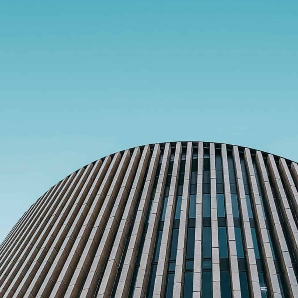 Low-angle view of a modern building with a curved facade, featuring vertical beige panels and dark windows against a clear blue sky.