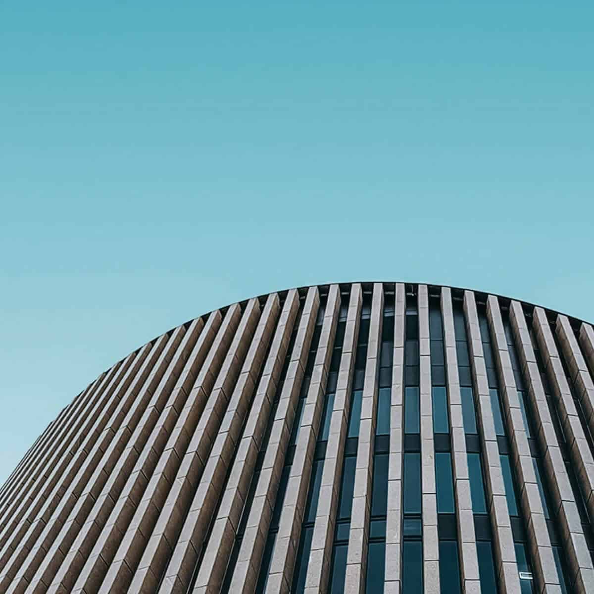 Low-angle view of a modern building with a curved facade, featuring vertical beige panels and dark windows against a clear blue sky.