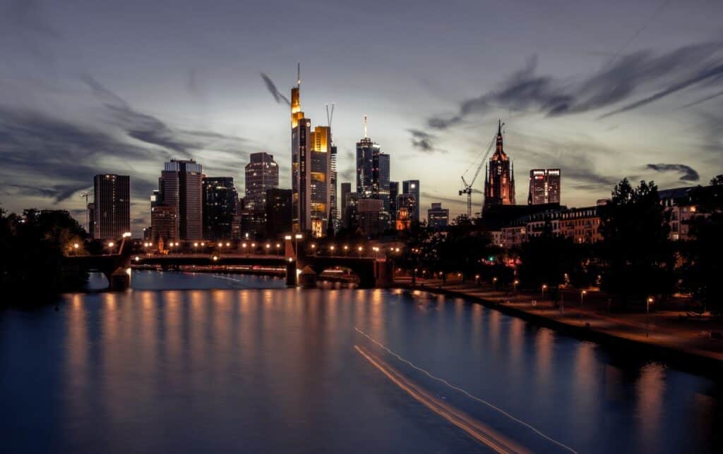 Night view of a city skyline with illuminated skyscrapers, a Gothic-style building, and a bridge over a river showing light trails.