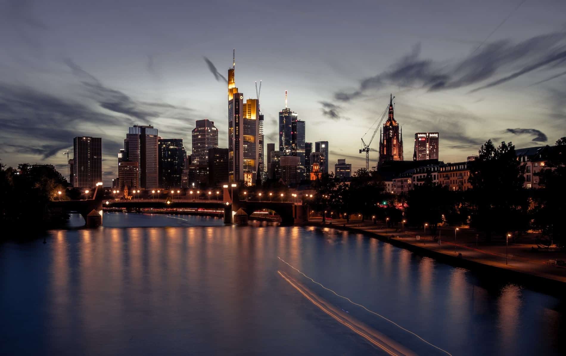 Night view of a city skyline with illuminated skyscrapers, a Gothic-style building, and a bridge over a river showing light trails.