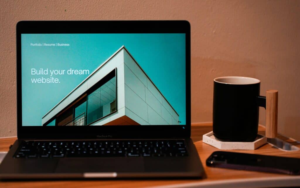 A MacBook Pro laptop on a wooden desk displays a website with a modern building and text 'Build your dream website.'. A black mug and smartphone are next to it.