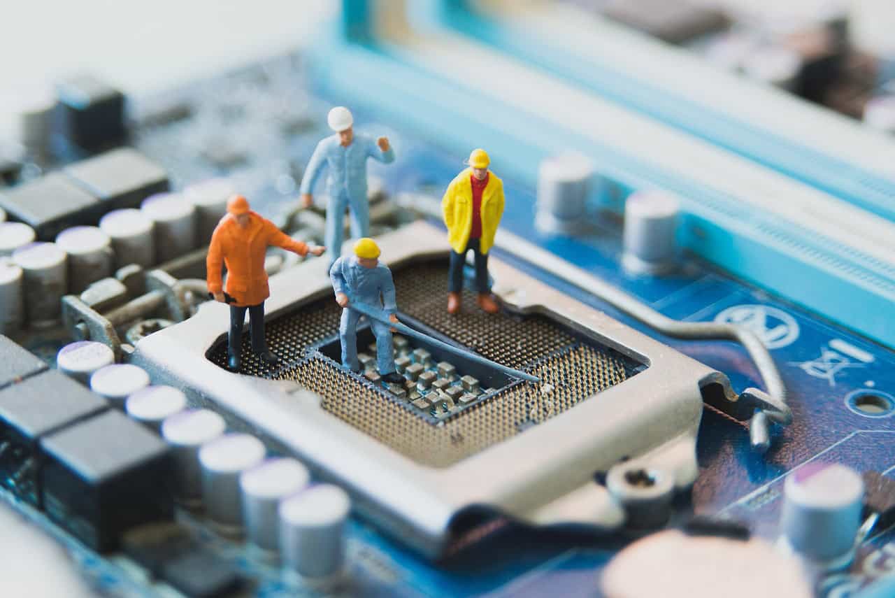 Four miniature worker figures on a blue computer motherboard, with one figure inside the CPU socket holding a tool, and others observing around it.