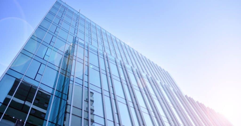 Low-angle view of a modern glass skyscraper with blue-tinted windows reflecting the sky and another building. A bright sun glare is visible in the bottom right.