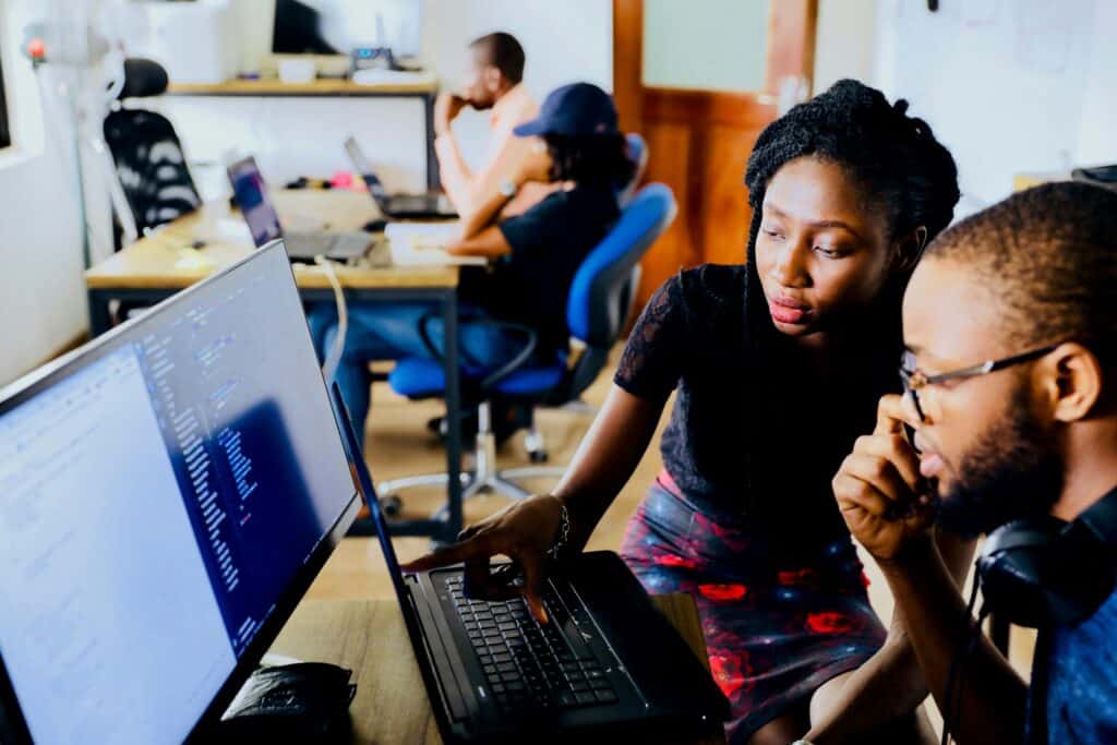 A woman points at a laptop screen displaying code, while a man in glasses looks on. Other people work in the blurred background.