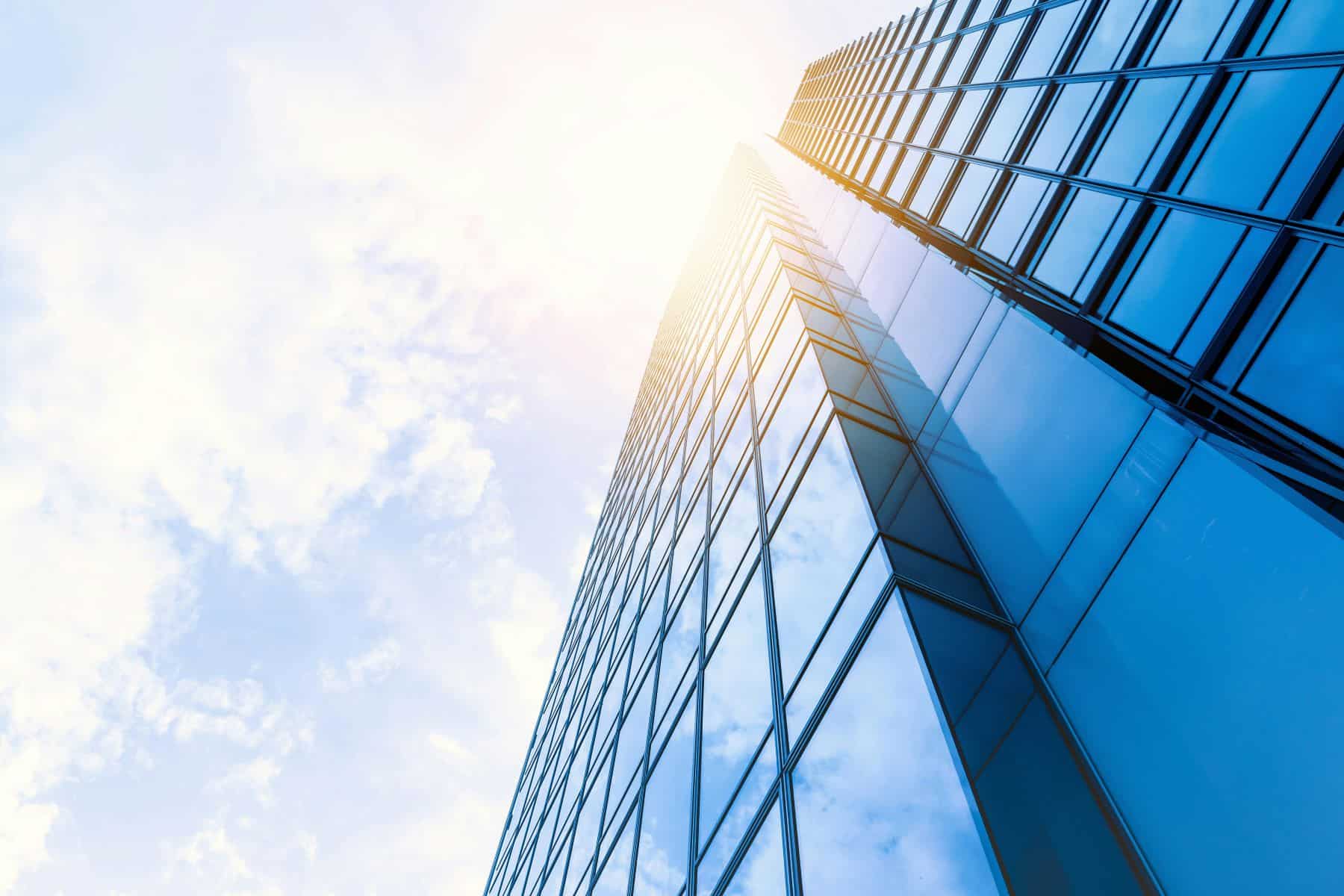 Low-angle view of a modern glass skyscraper with blue-tinted windows, reflecting a bright, cloudy sky with sun glare.