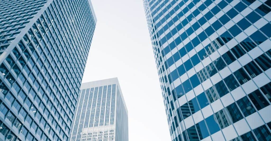 Low-angle view of three modern skyscrapers with glass facades against a bright sky. Two large buildings flank a smaller one in the background.