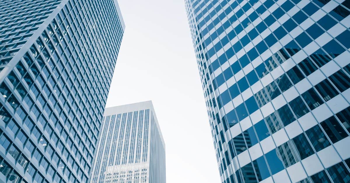 Low-angle view of three modern skyscrapers with glass facades against a bright sky. Two large buildings flank a smaller one in the background.