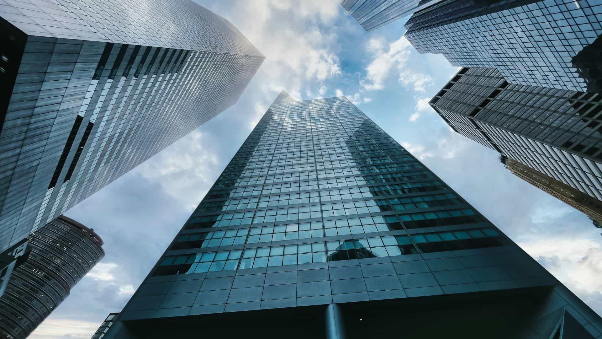 Low-angle view of several modern glass skyscrapers reaching towards a cloudy blue sky.