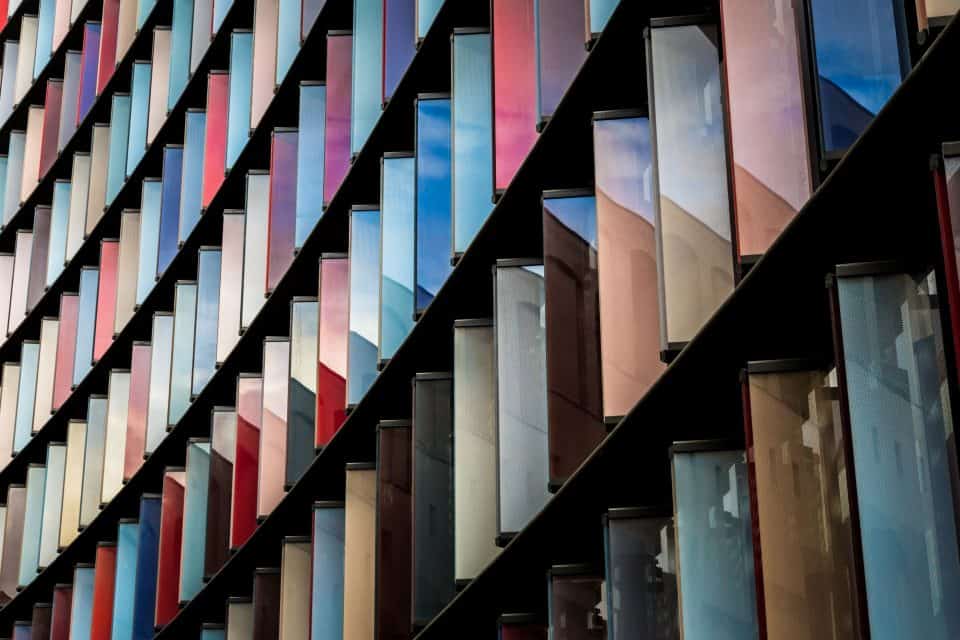 Close-up of a modern building facade with diagonal rows of colorful, reflective rectangular panels in various shades of blue, red, pink, and brown.