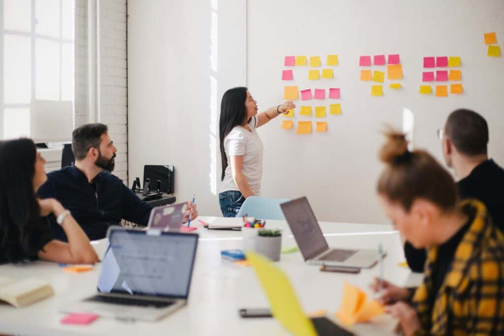 A woman presents ideas on a wall covered with colorful sticky notes to a group of colleagues in a bright office.