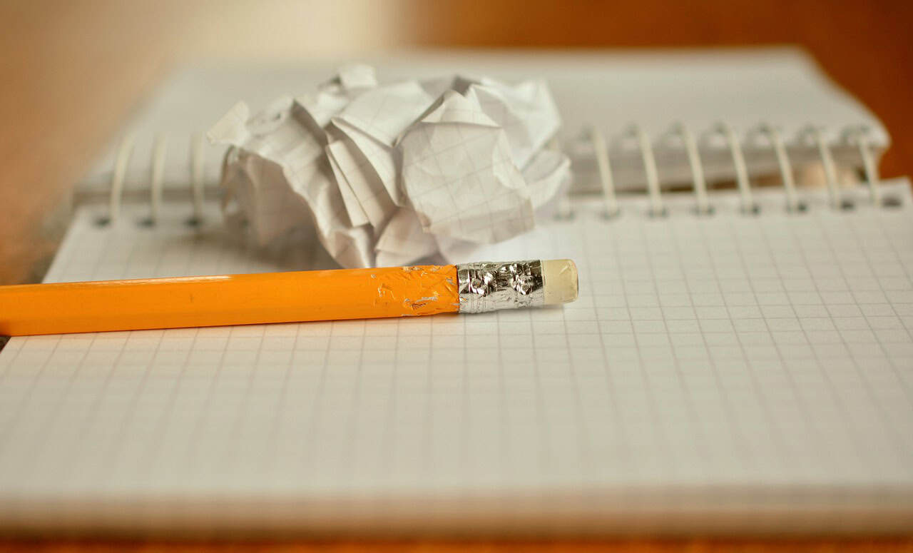 A yellow pencil with a worn eraser, a crumpled piece of grid paper, and an open spiral notebook on a desk.