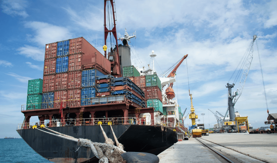 A large container ship docked at a port, laden with red, blue, and green shipping containers, with cranes and port infrastructure visible under a blue sky.