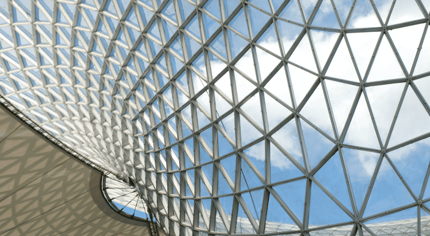 Upward view of a curved glass and metal geodesic dome with triangular panels, showing blue sky and white clouds through it. A white fabric structure below has geometric shadows.