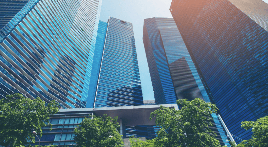 Low-angle view of multiple modern skyscrapers with blue glass facades and green trees in the foreground under a bright sky.