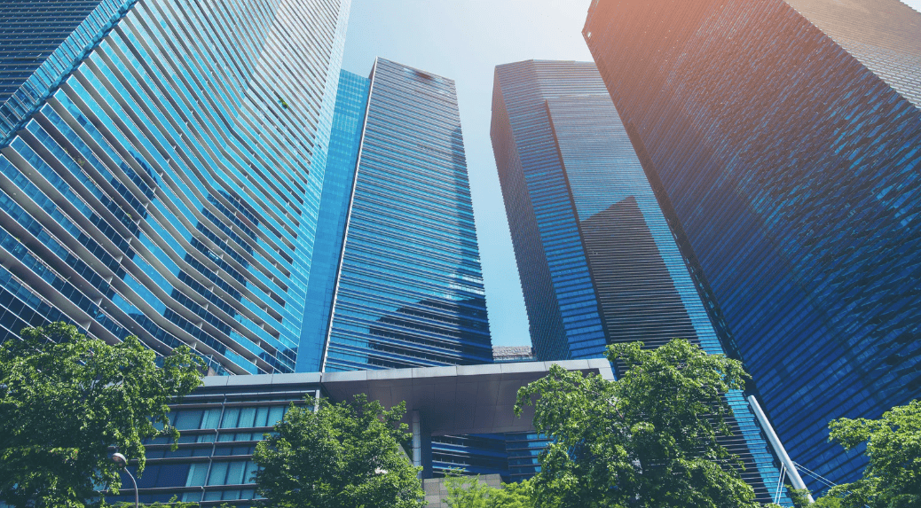 Low-angle view of multiple modern skyscrapers with blue glass facades and green trees in the foreground under a bright sky.