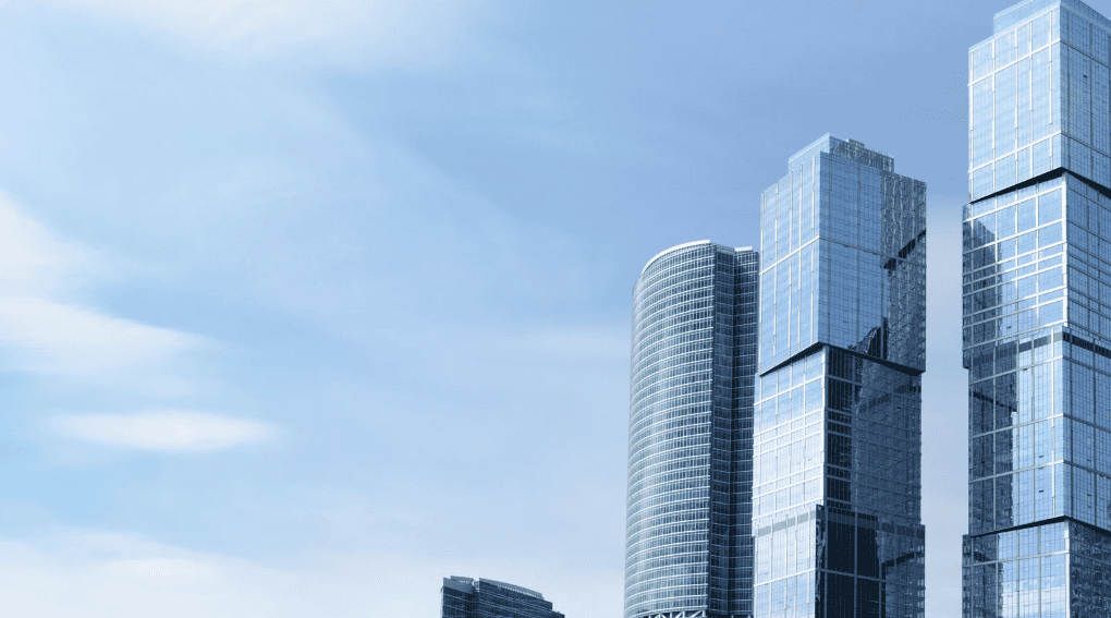 Modern glass skyscrapers against a light blue sky with faint clouds. Buildings are rectangular and curved, reflecting the sky.