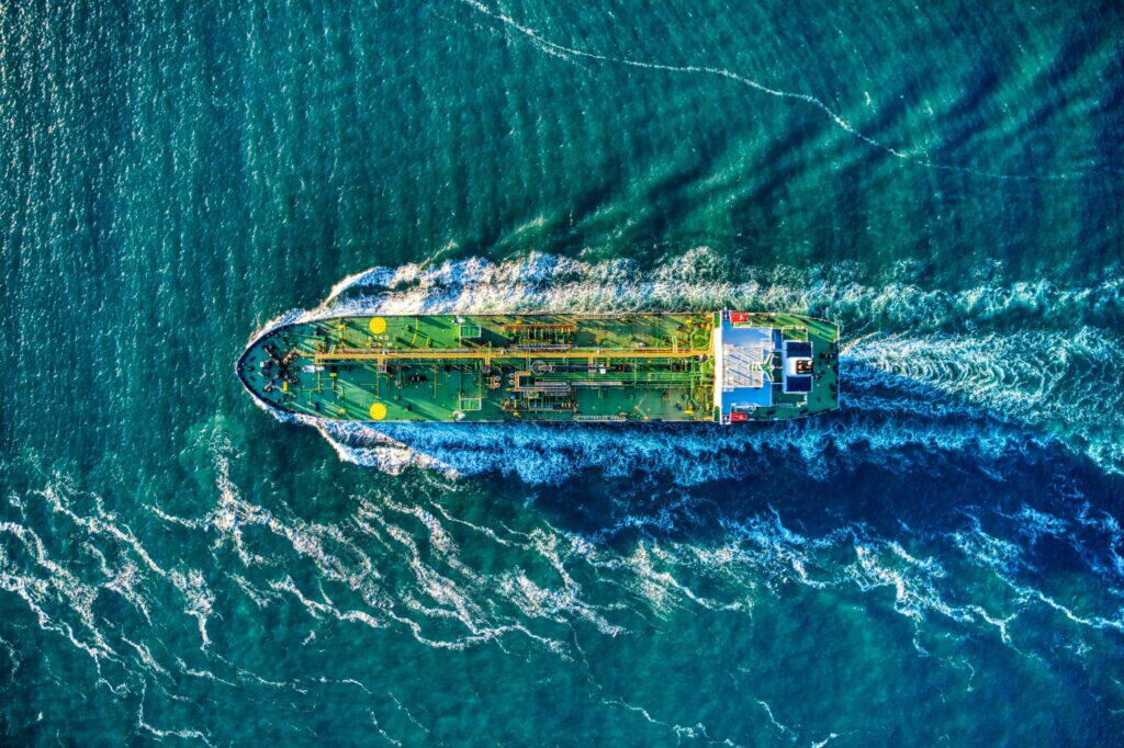 Aerial top-down view of a green cargo ship with a white superstructure sailing through blue-green ocean water, leaving a white foamy wake.
