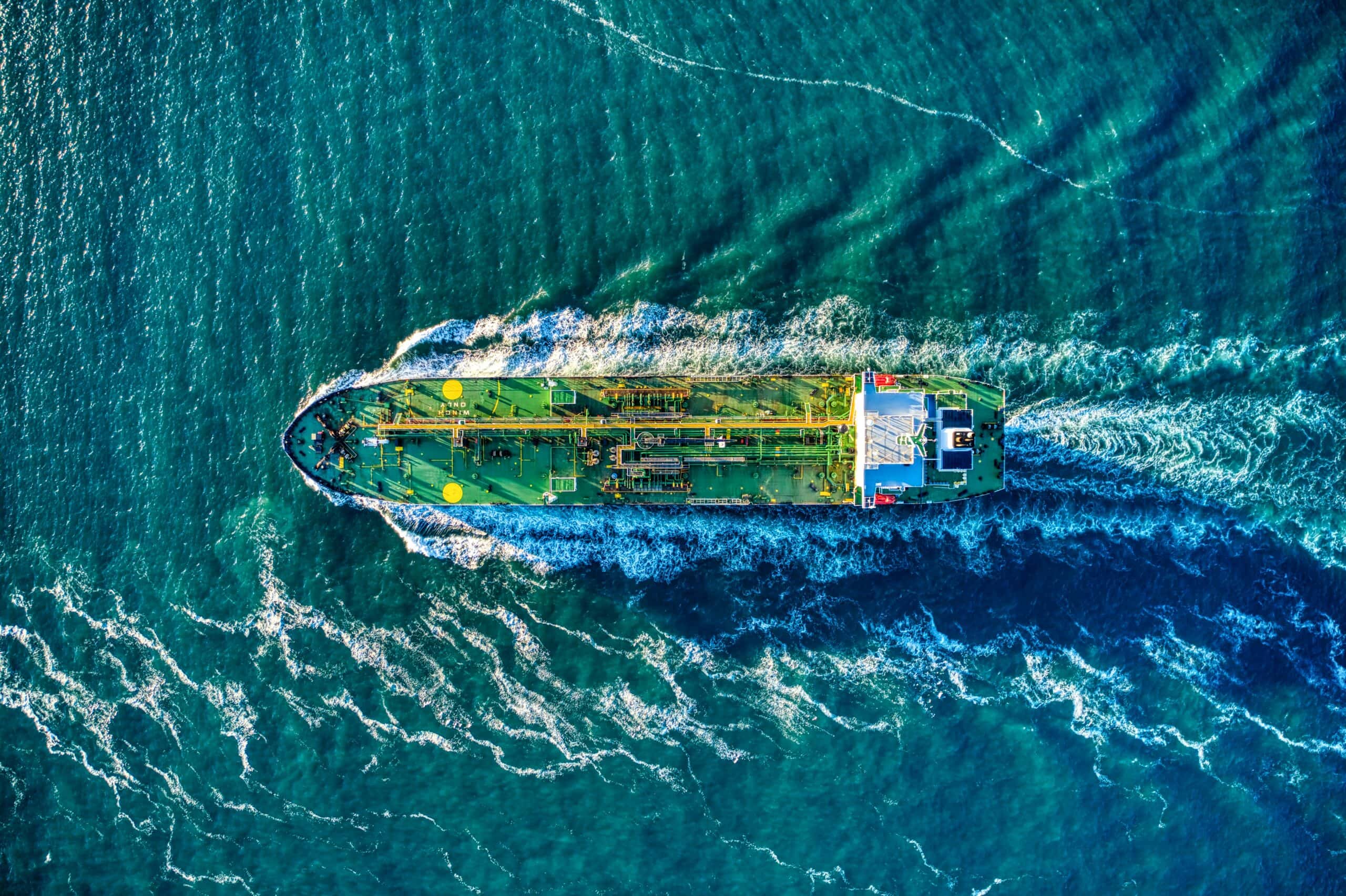 Aerial top-down view of a green cargo ship with a white superstructure sailing through blue-green ocean water, leaving a white foamy wake.