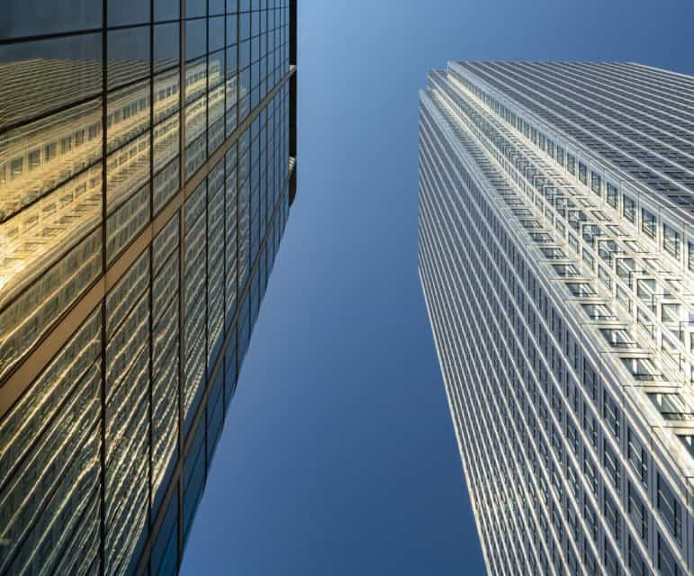 Two tall skyscrapers, one glass and one light-colored, viewed from below against a clear blue sky. The glass building reflects a golden light.