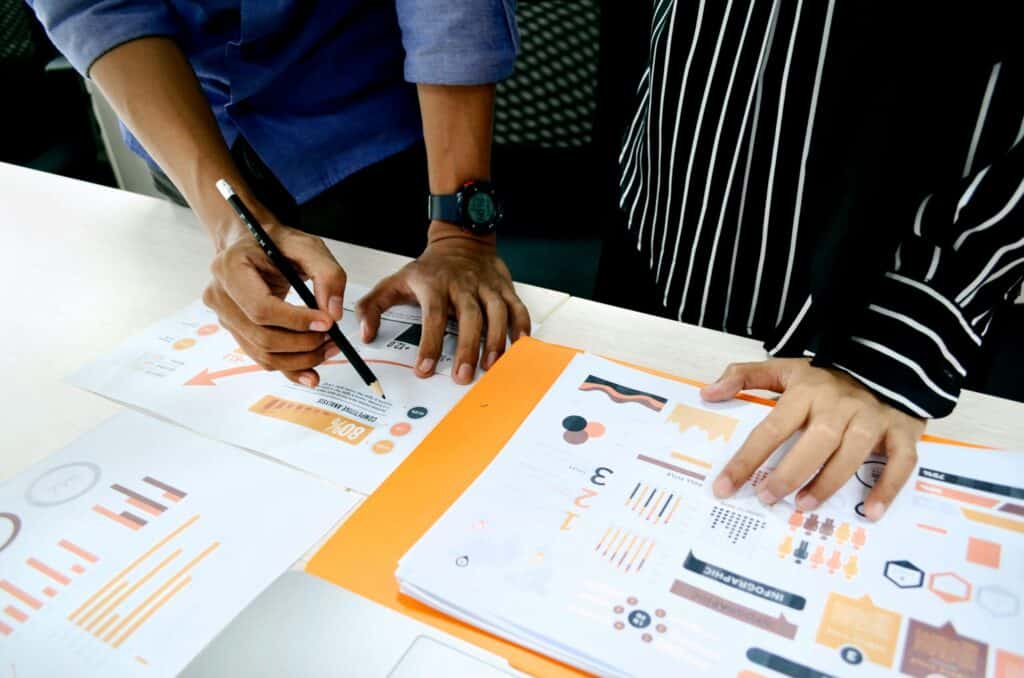 Two people's hands reviewing business documents with charts, graphs, and text on a desk.