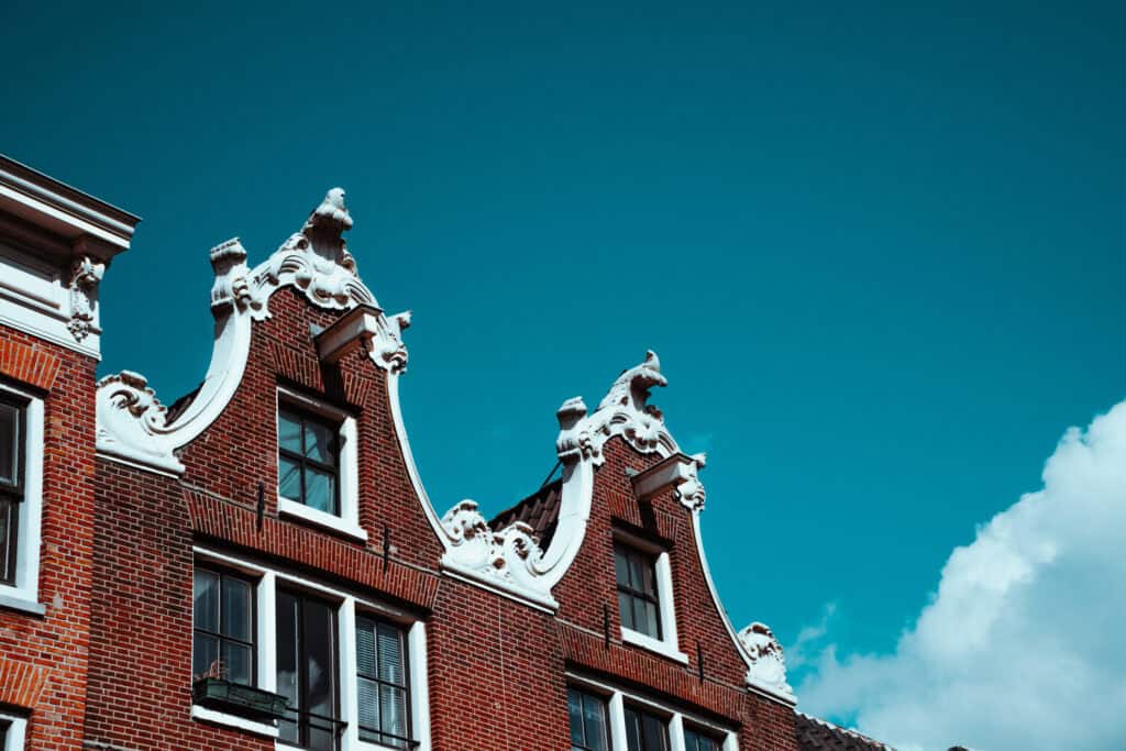 Upper part of red brick gabled buildings with ornate white decorative elements under a clear blue sky with a white cloud.