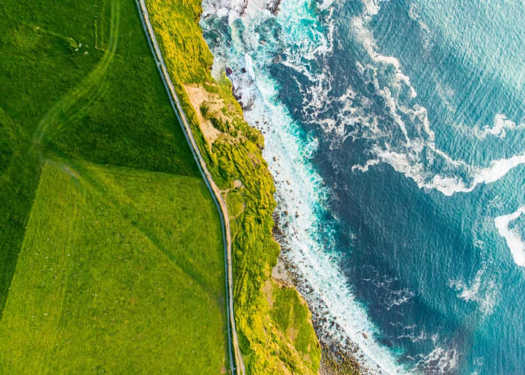 Aerial view of a green grassy field and a dirt path running alongside a rocky, green-covered coastline, with deep blue ocean waves crashing against the shore.
