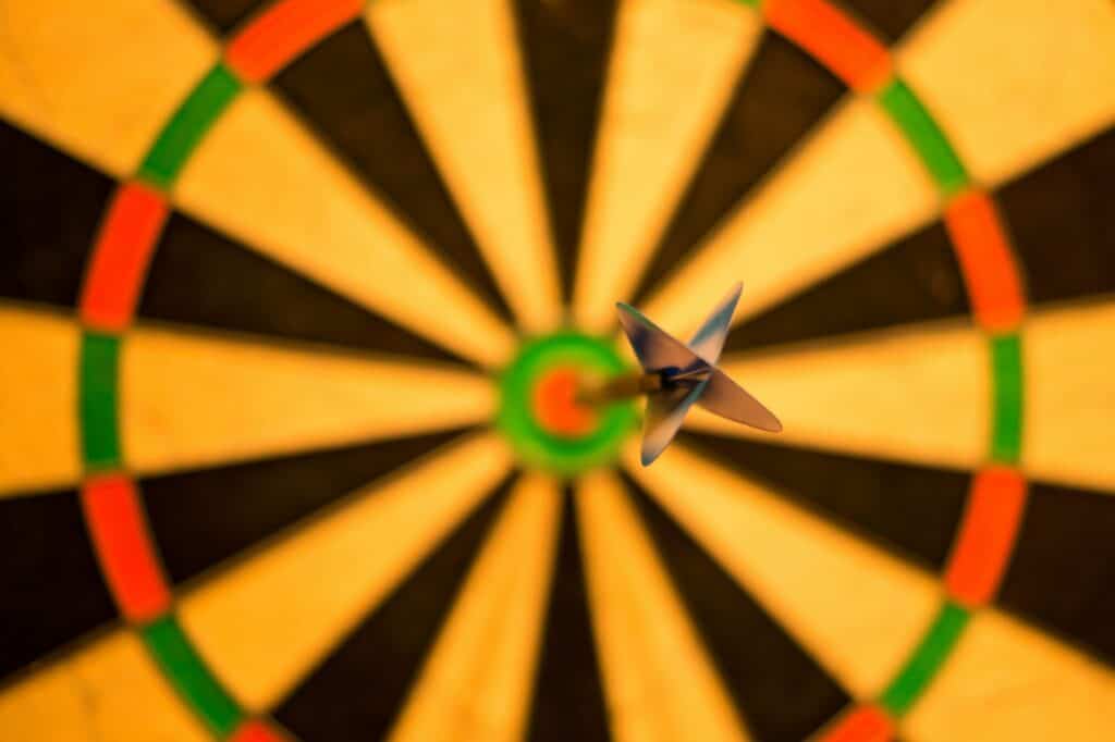 A dart with light blue fletching stuck directly in the red bullseye of a yellow, black, green, and red dartboard.
