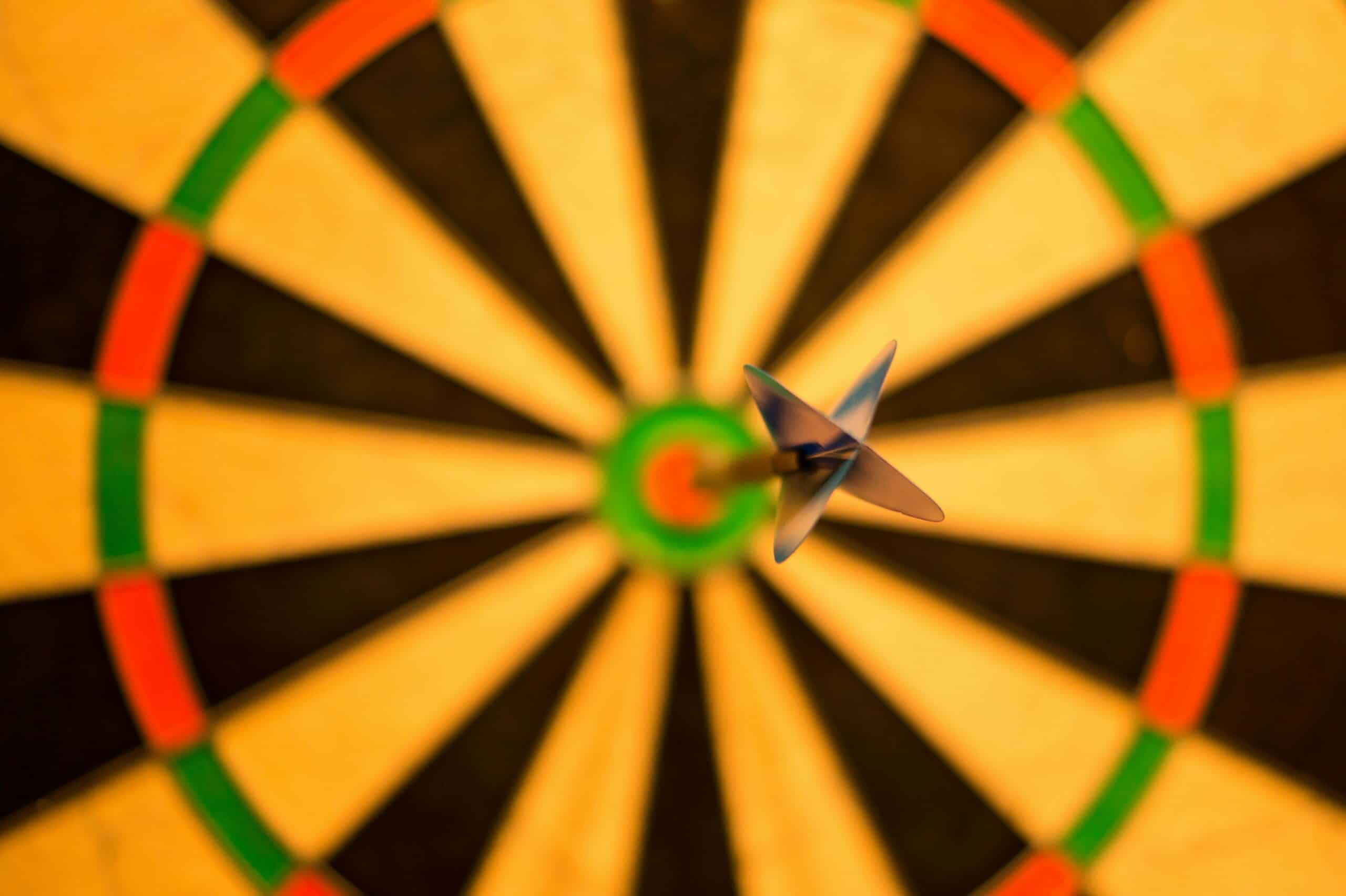 A dart with light blue fletching stuck directly in the red bullseye of a yellow, black, green, and red dartboard.