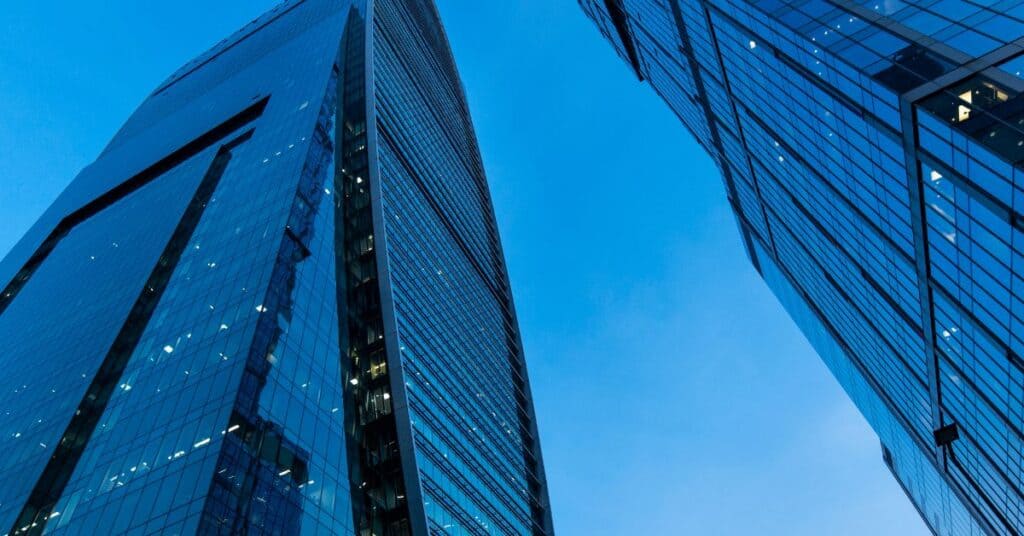 Two modern glass skyscrapers with blue reflective facades against a clear blue sky, viewed from below.