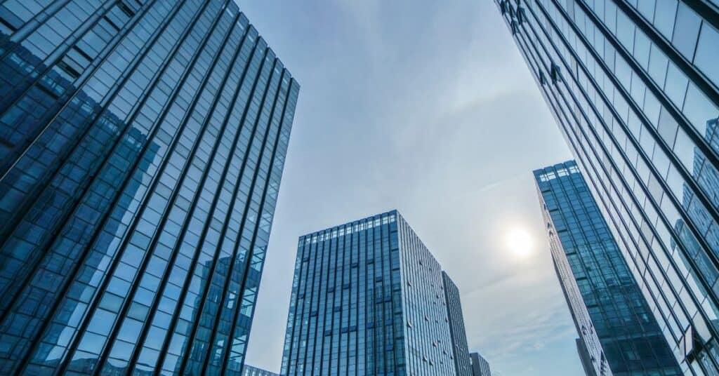 Low-angle view of multiple blue glass skyscrapers under a bright sky with the sun visible.