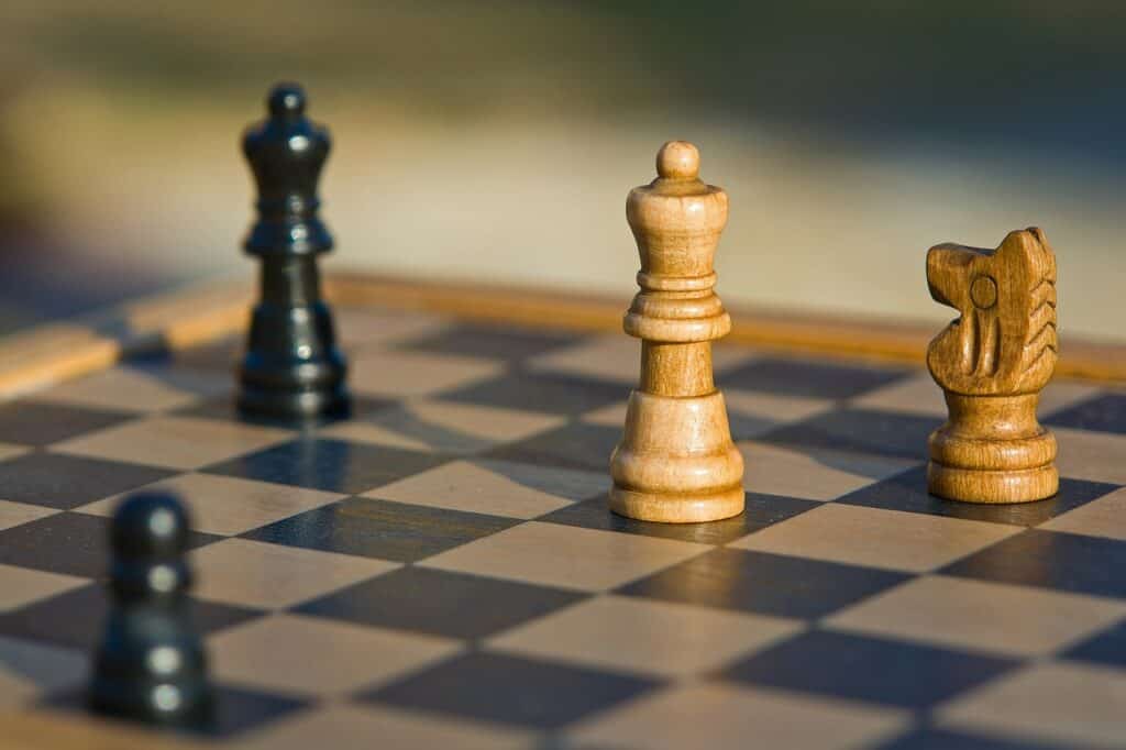 A close-up of a wooden chessboard with a white queen and knight in focus, and blurred black queen and pawn.