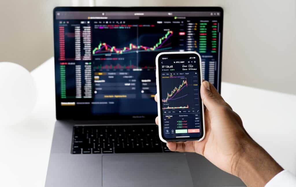 A person's hand holds a smartphone displaying a cryptocurrency trading chart, with a laptop showing similar financial data in the background.
