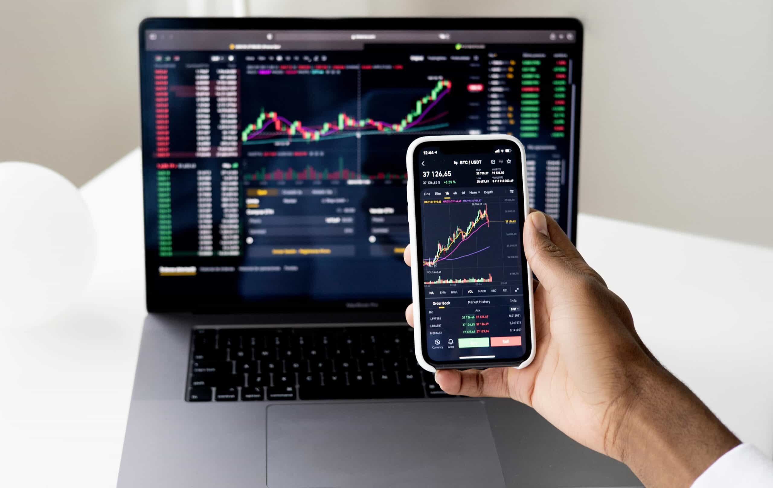 A person's hand holds a smartphone displaying a cryptocurrency trading chart, with a laptop showing similar financial data in the background.