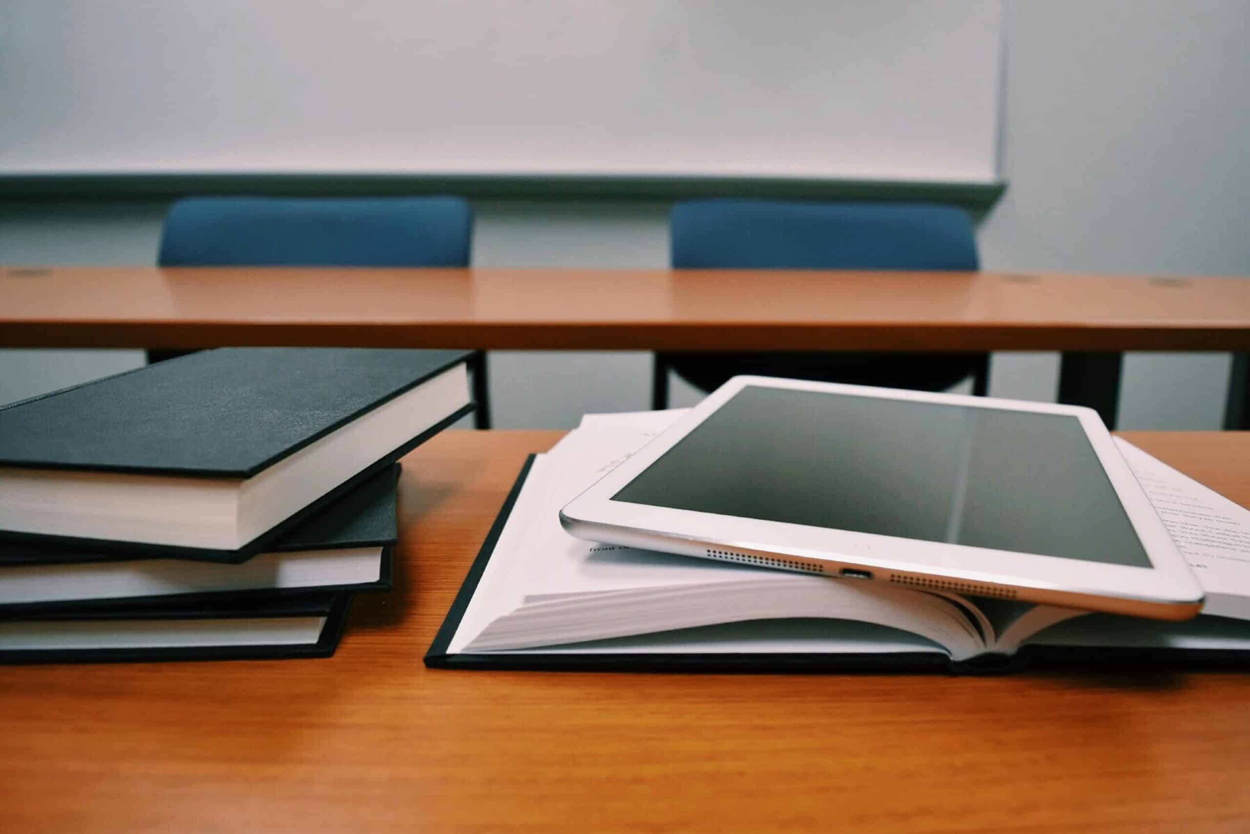 A stack of black-covered books and a silver tablet on an open book sit on a wooden desk in a classroom.