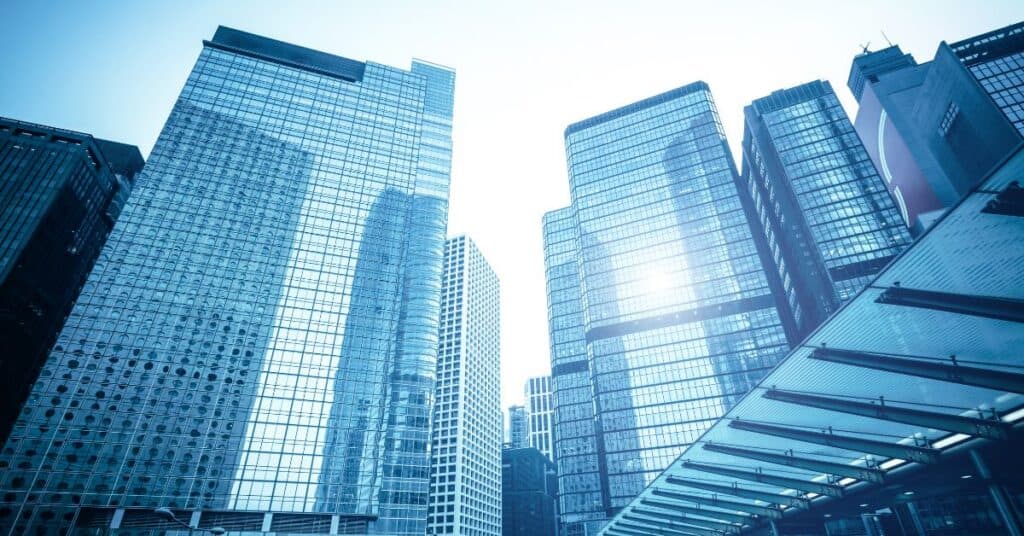 Low-angle view of multiple modern glass skyscrapers under a bright sky with sun glare.