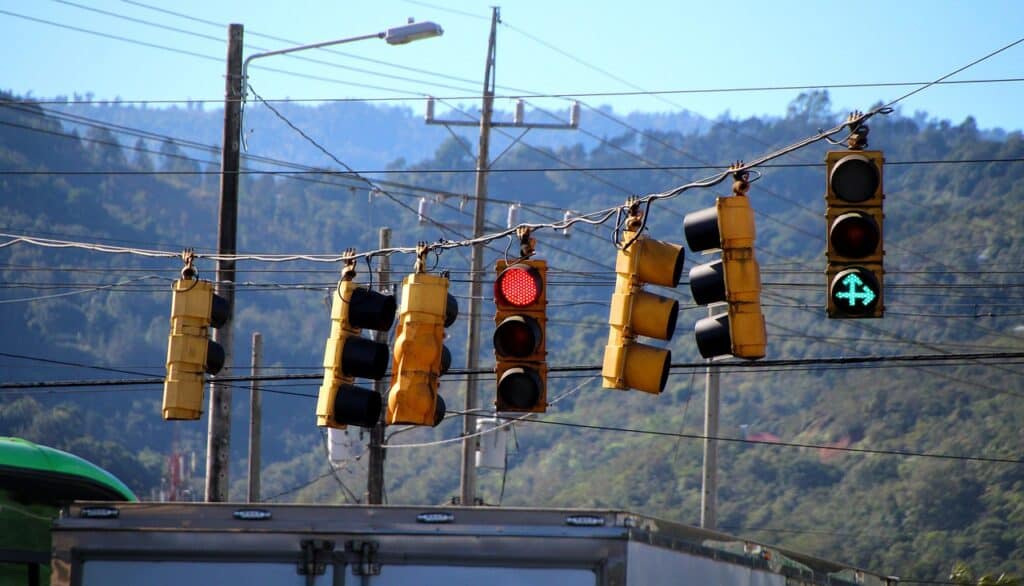 Multiple yellow traffic lights and power lines hang over a street, with a red light and a green right arrow visible. Distant green mountains are in the background.