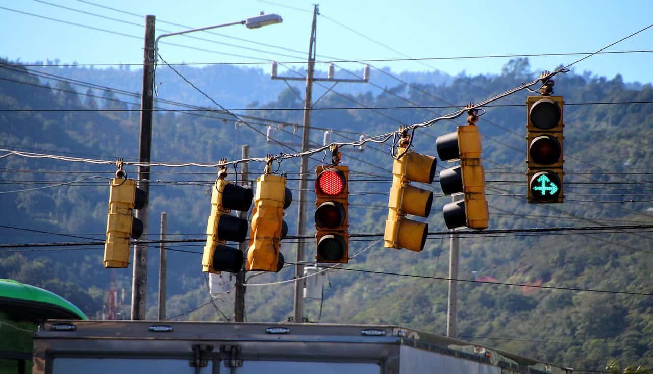 Multiple yellow traffic lights and power lines hang over a street, with a red light and a green right arrow visible. Distant green mountains are in the background.