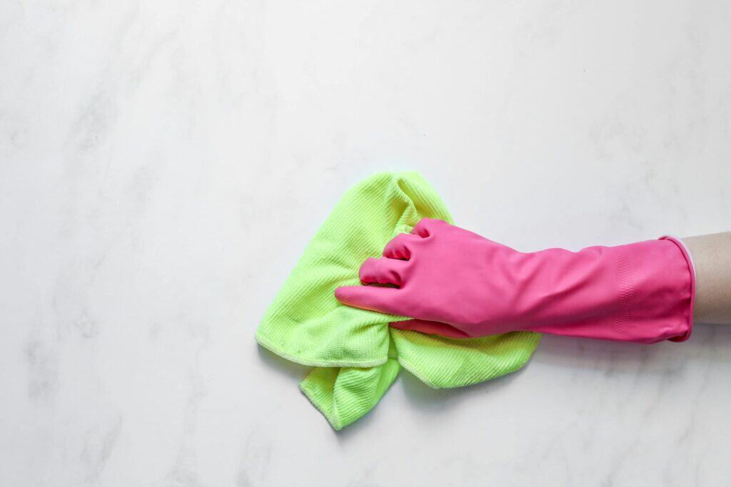 A hand in a pink rubber glove holds a green microfiber cleaning cloth against a white marble background.