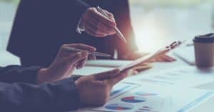 Close-up of two people's hands with pens, reviewing business documents with charts and graphs on a table.