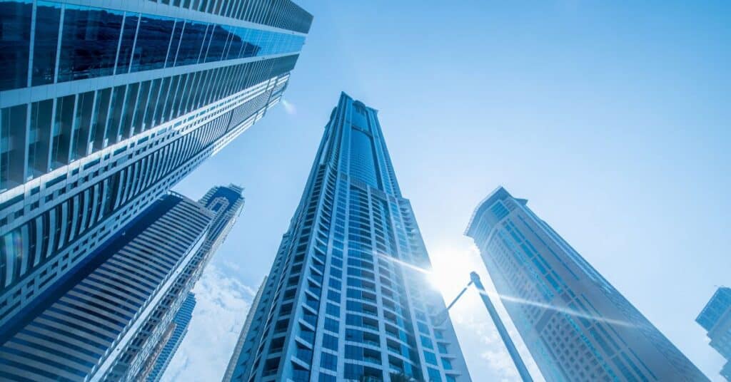 Low-angle view of multiple modern glass skyscrapers reaching into a clear blue sky, with a bright sun flare visible.